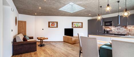 A modern living space with a wooden floor, a sofa, a dining table with chairs, a TV, and kitchen cabinets.