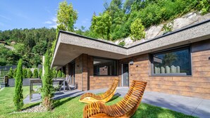 Ein modernes Haus mit geneigtem Dach, einer Terrasse mit zwei Holzliegen und Blick auf einen bewaldeten Hügel.