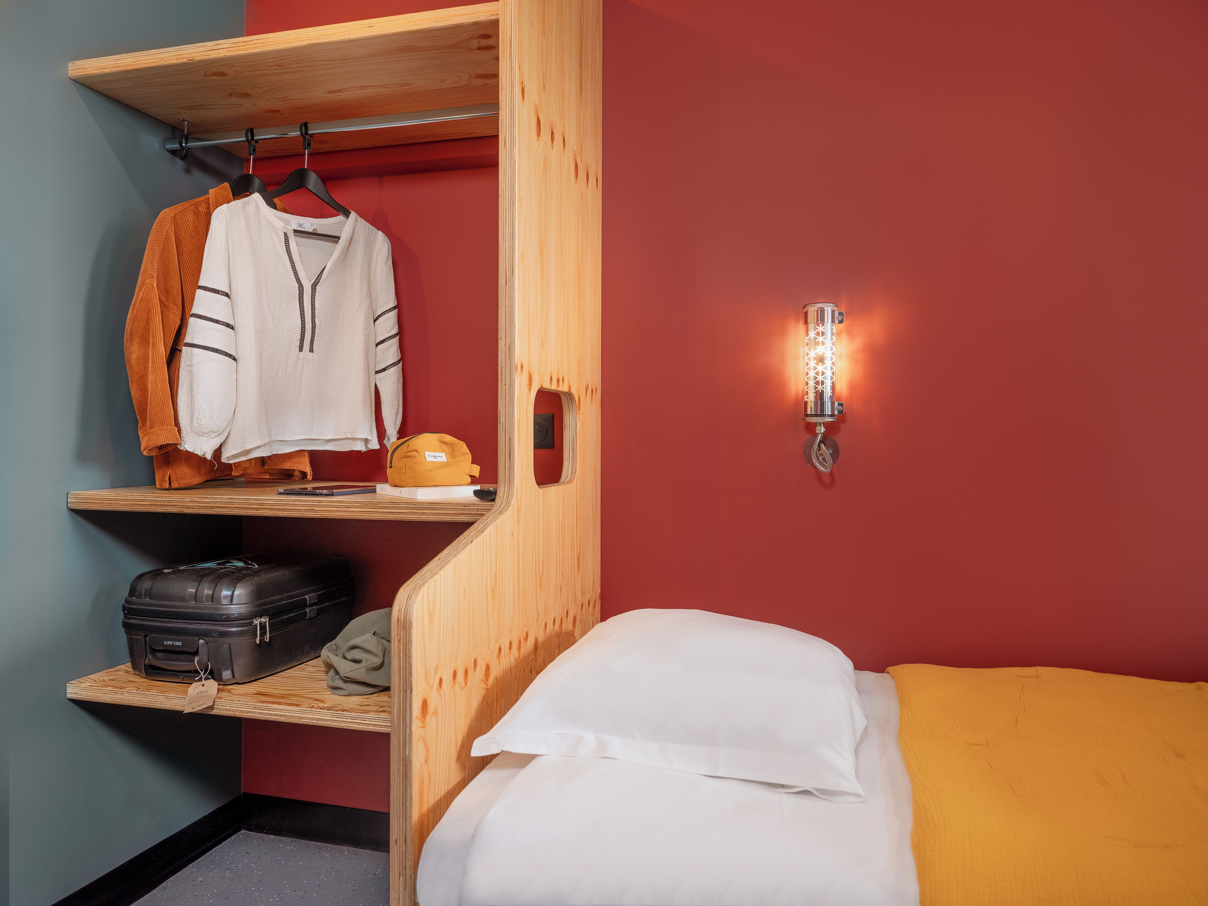 A compact hotel room with a bed, a wooden shelf with a hanging garment, a suitcase, and a wall-mounted light fixture.