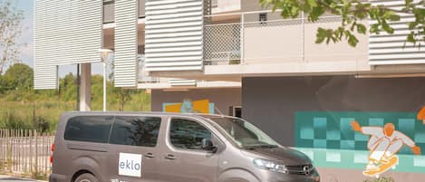 A gray van parked in front of a building with a blue 'eklo' sign, a mural with a figure, and a stone wall.
