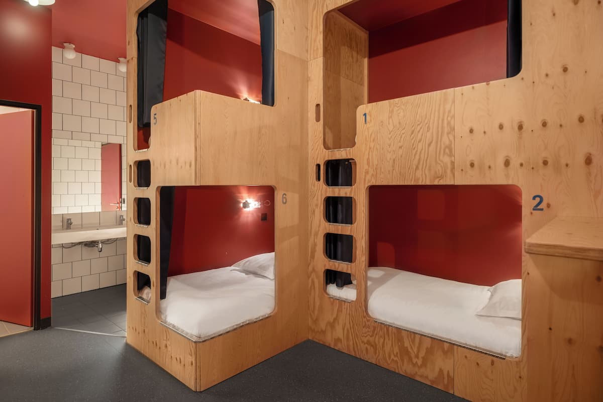 Una habitación con dos literas, cada una con una ventana pequeña y un espejo. Se ve un lavamanos y un inodoro al fondo.