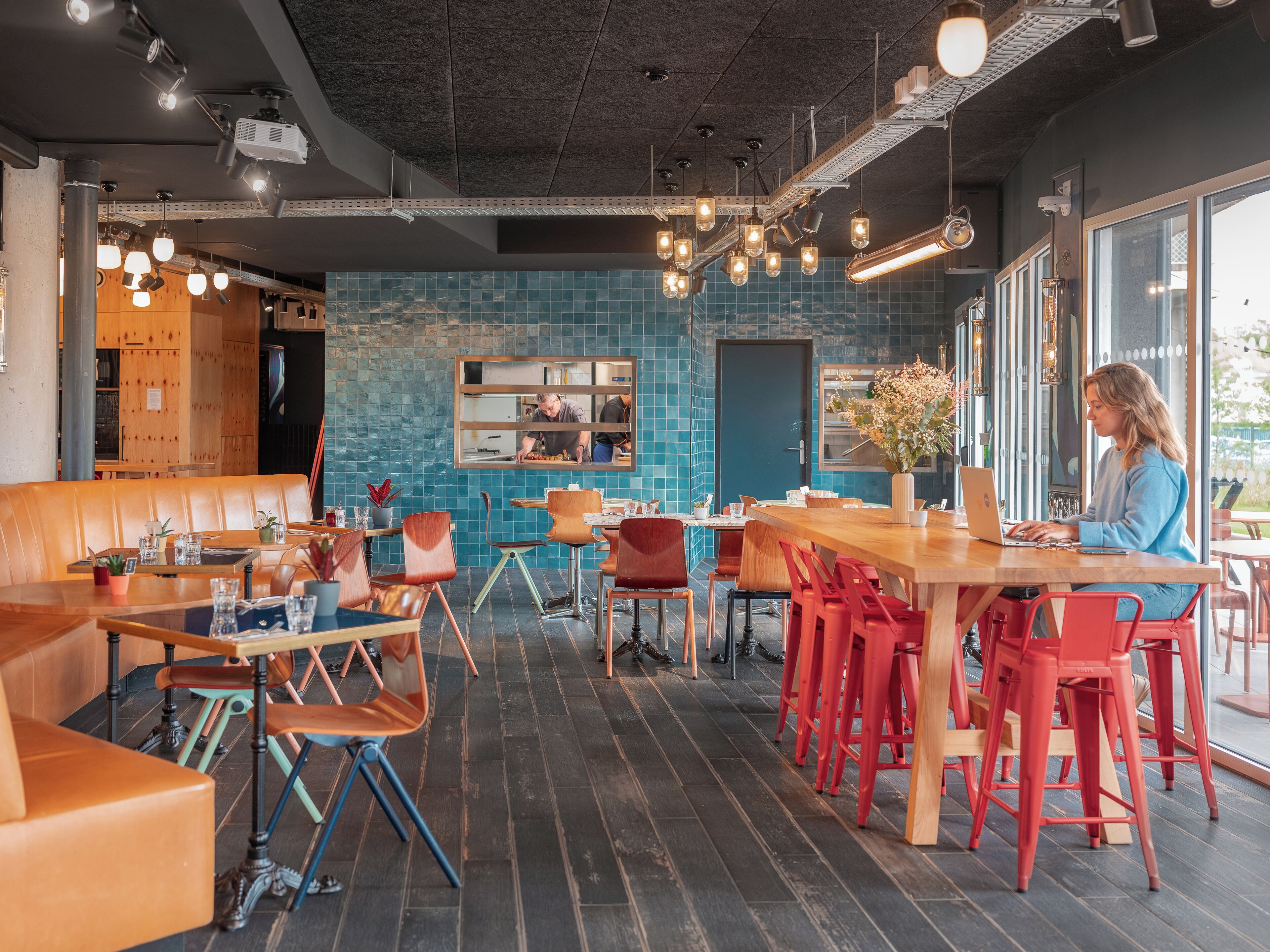 A modern cafe with wooden tables and red chairs, a blue-tiled wall, and a large window.