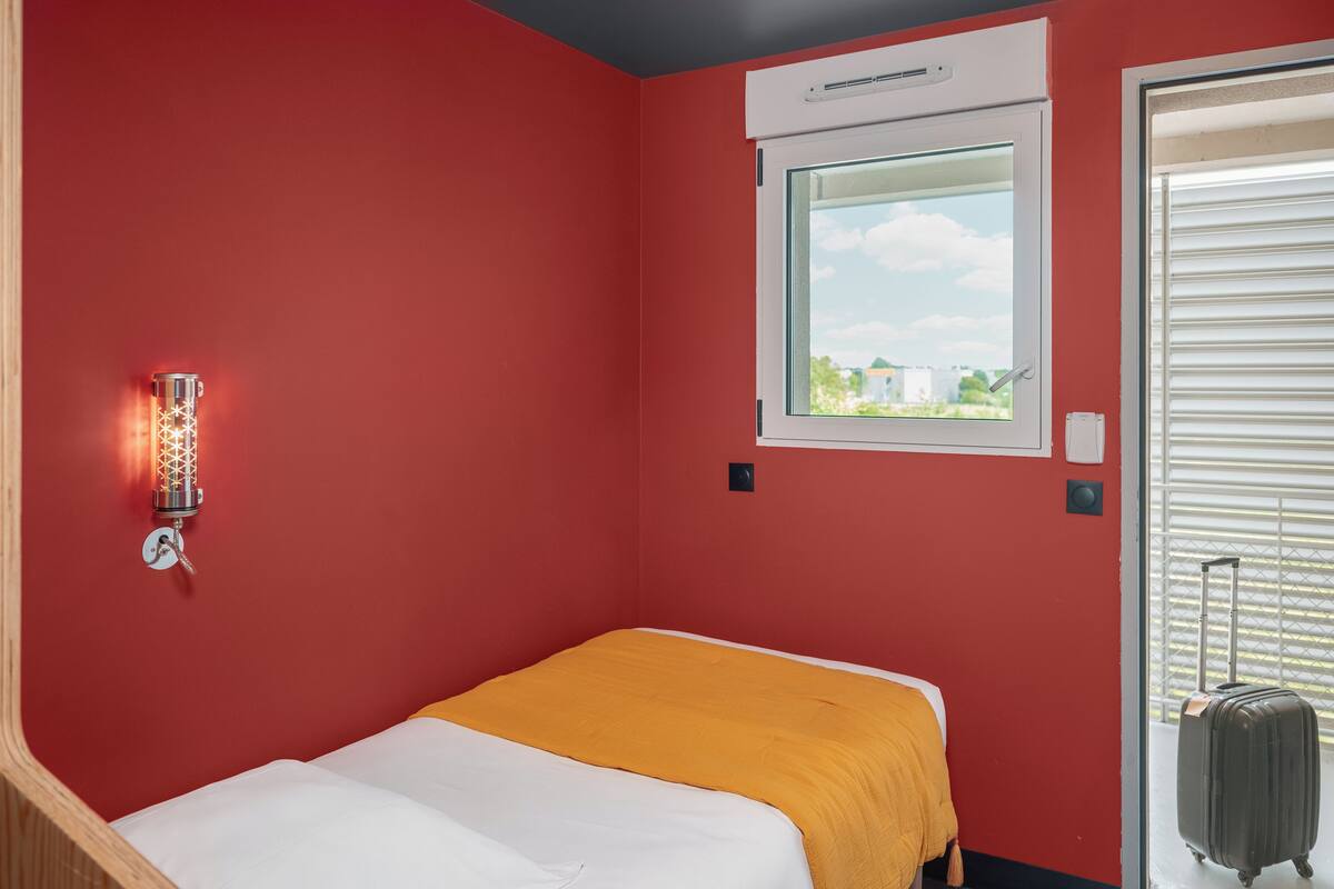 Habitación pequeña con cama individual, pared roja, ventana con vistas, luz fijada en la pared y una maleta cerca de la puerta.
