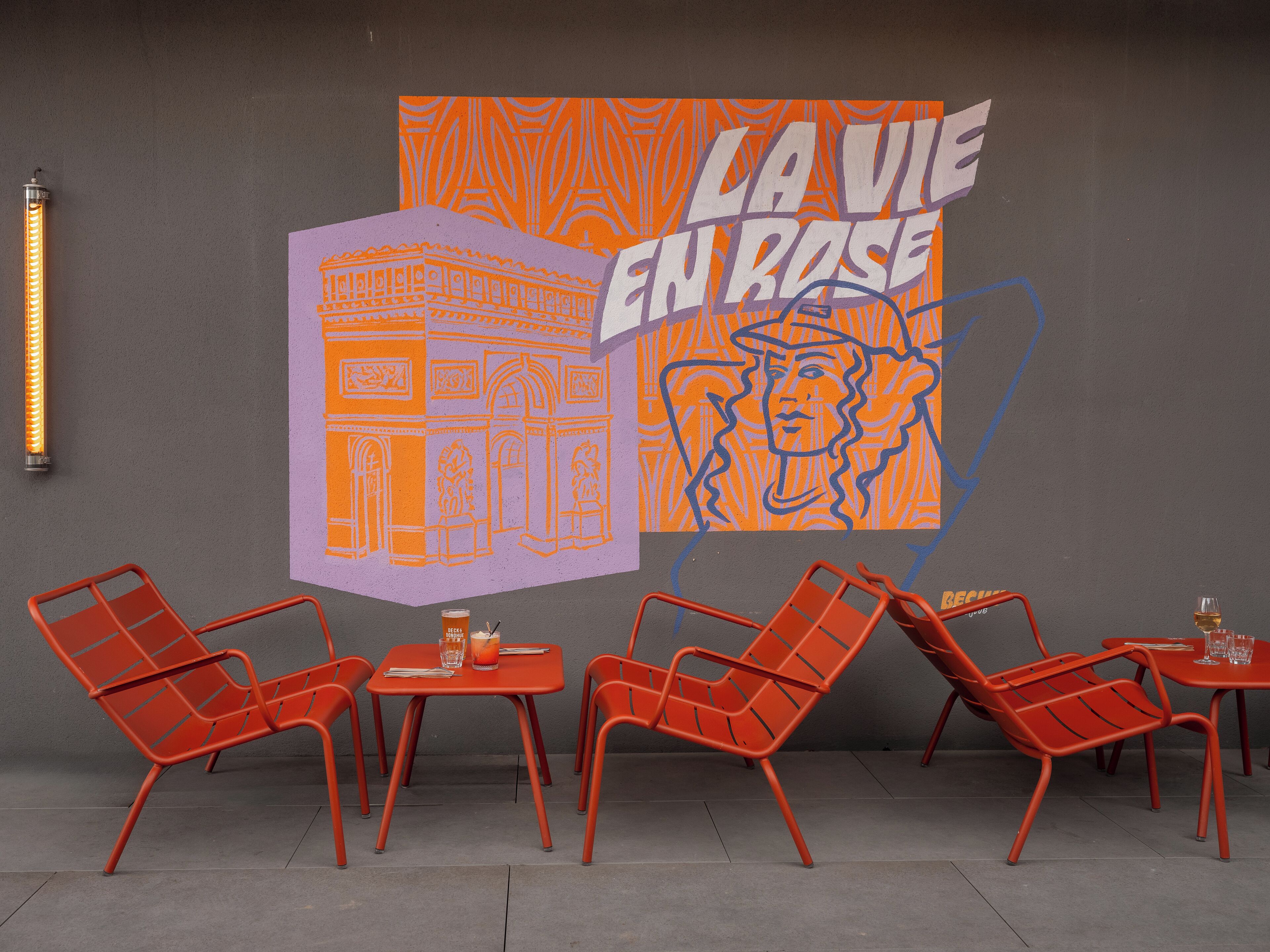 A modern outdoor seating area with red metal chairs and tables, a mural with 'LA VIE EN ROSE' text, and a tall, cylindrical light fixture.