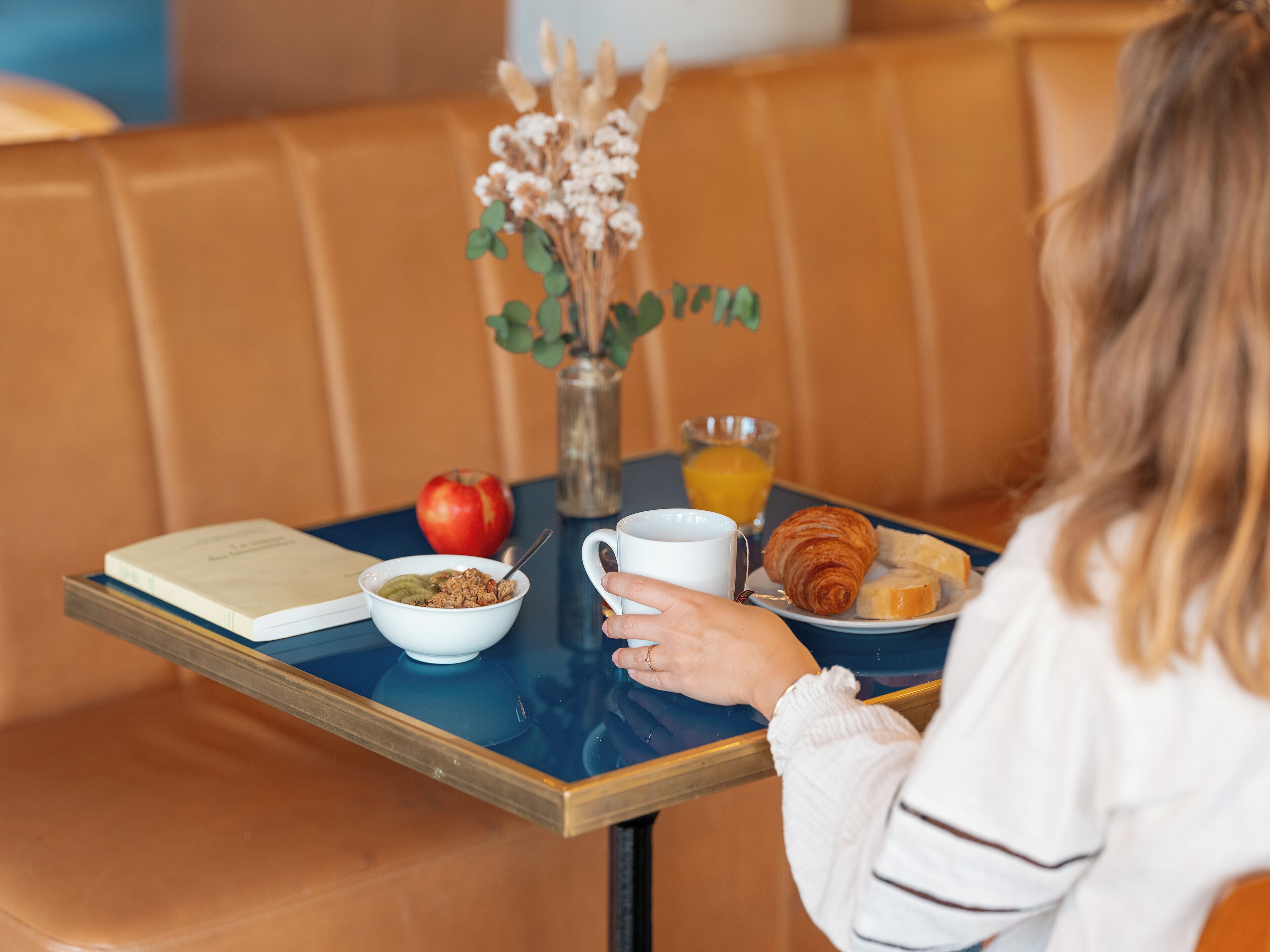 A breakfast table with a book, bowl of cereal, apple, glass of orange juice, coffee cup, croissant, and a vase with dried flowers.