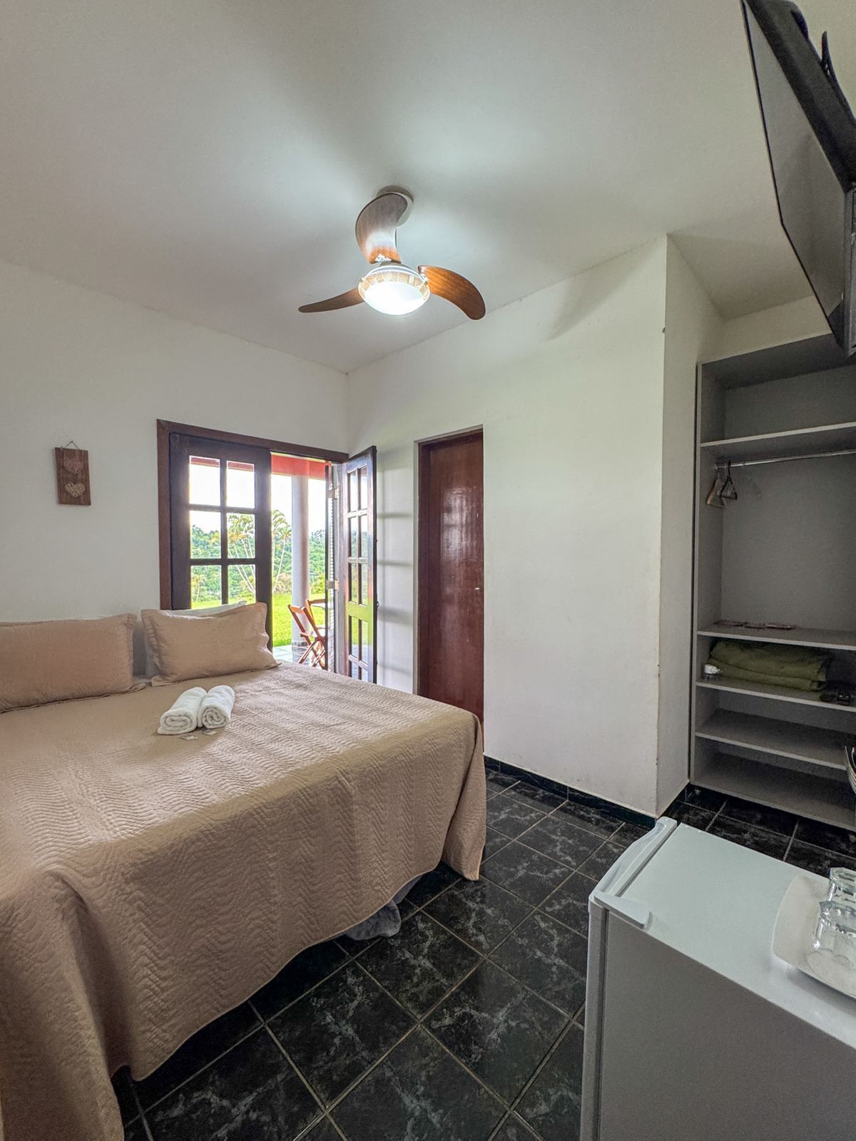 Quarto com cama, ventilador de teto, armário branco e vista para o exterior.
