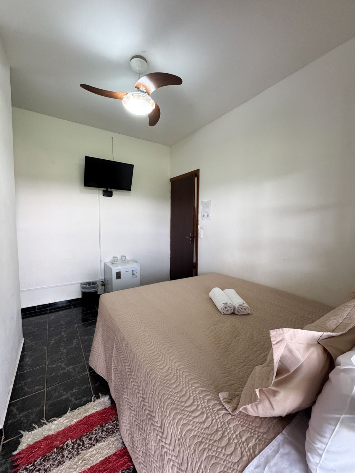A hotel room with a bed, a ceiling fan, a television, a small refrigerator, and a door.