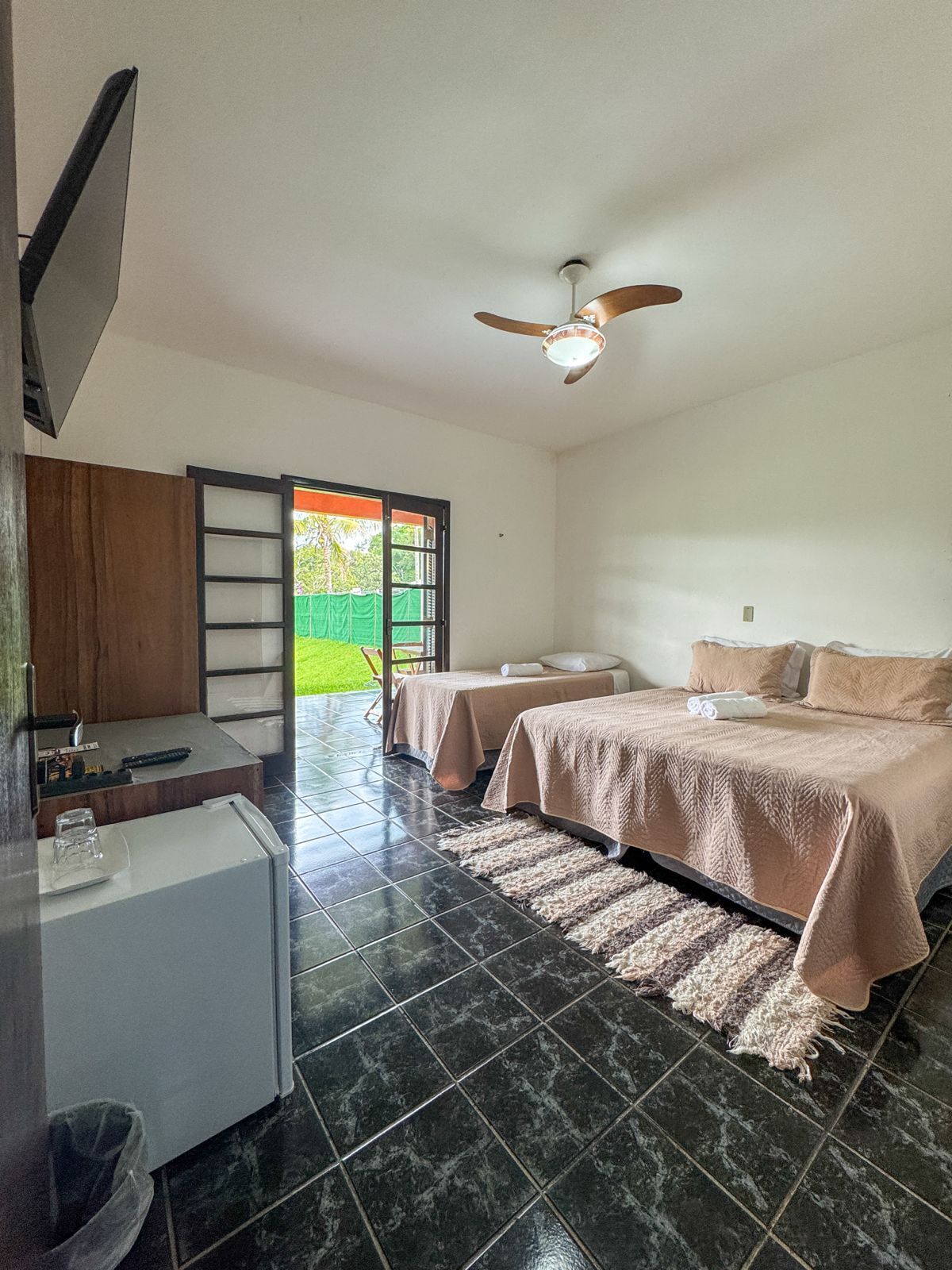 A hotel room with a bed, a desk, a chair, a ceiling fan, and a view of the outdoors.