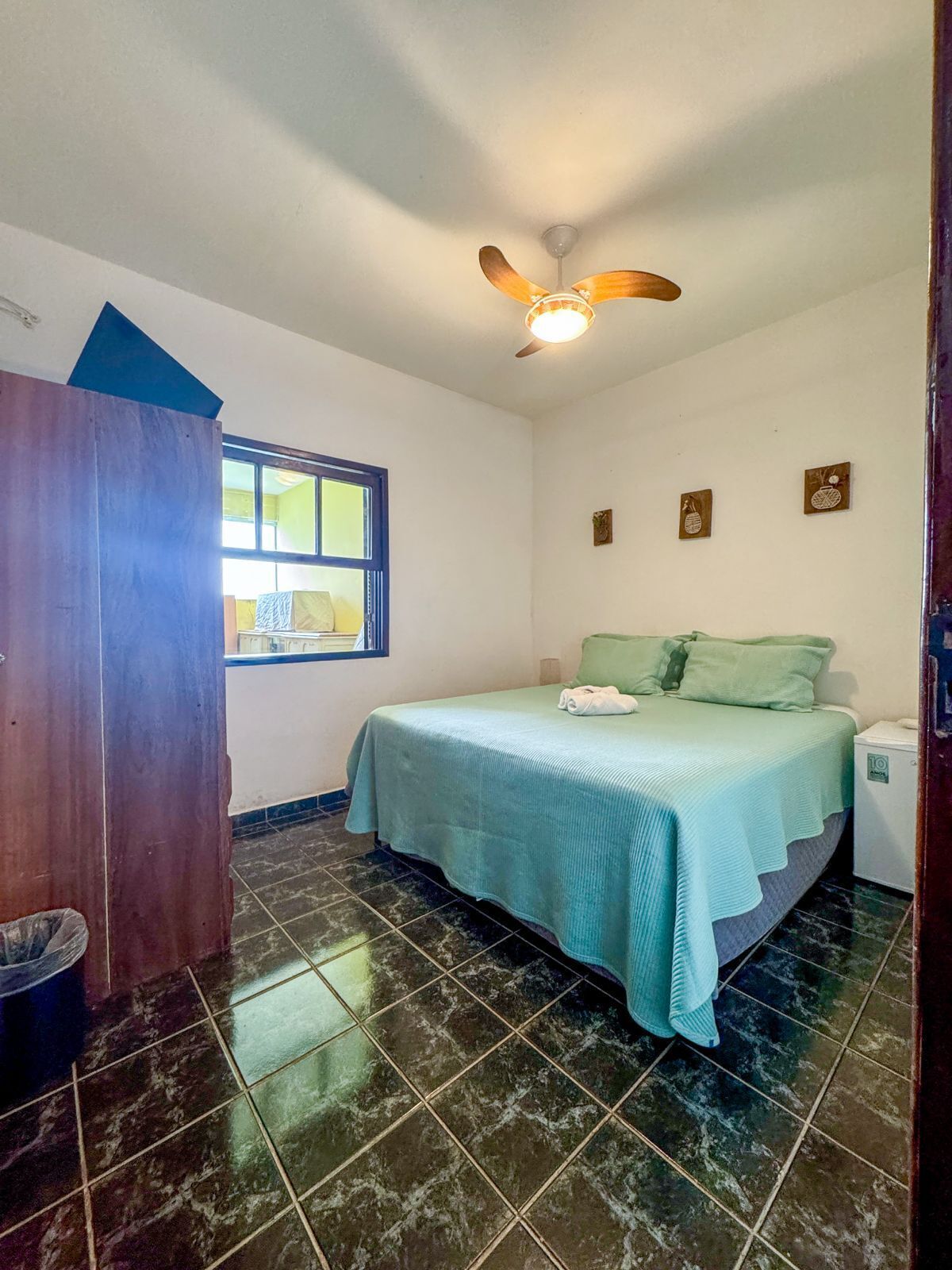 Quarto com cama, ventoinha no teto, janela e uma mesinha de cabeceira.