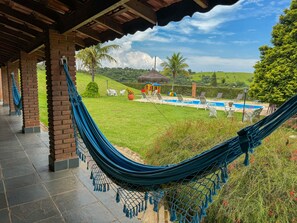 A hammock under a wooden pergola overlooking a pool area with lounge chairs and a thatched-roof structure. - Pousada Espaço do Sol (Aracariguama)