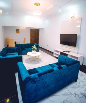 A modern living room with a blue sofa set, a white coffee table, and a flat-screen TV on a white stand. - Mconyx Apartment (Lagos)
