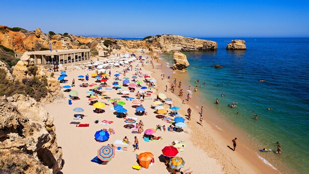 Une plage parsemée de nombreux parasols colorés, bordée d’une eau d’un bleu limpide et de falaises rocheuses.