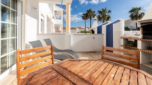 Une table et des chaises en bois, disposées dans un espace extérieur ensoleillé, avec vue sur des palmiers et un bâtiment aux touches de bleu.