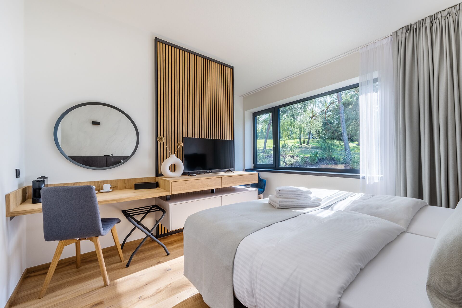 A modern hotel room with a large bed, a desk with a computer, a chair, a round mirror, and a window with a view of greenery.