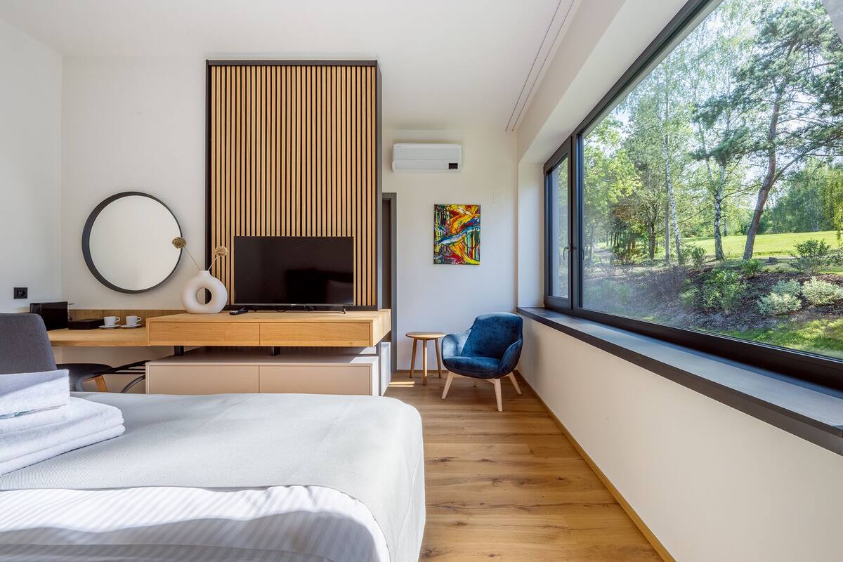 Moderni hotellihuone, jossa on suuri sänky, työpöytä, jossa on litteänäyttöinen televisio, pieni sivupöytä, sininen nojatuoli ja suuri ikkuna, josta avautuu näkymä metsään.