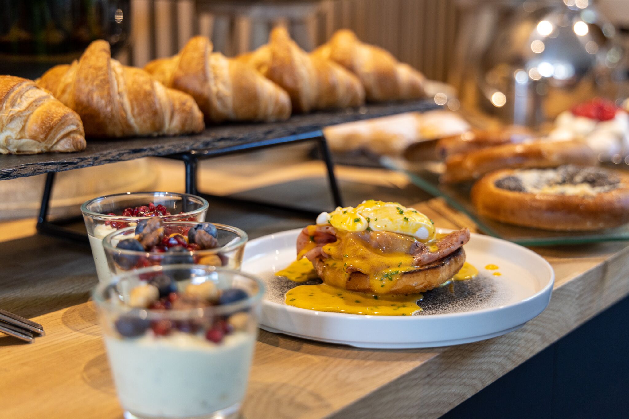 A breakfast buffet with croissants, a plate of eggs Benedict, and glasses of yogurt with berries.