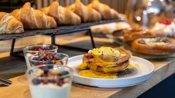 A breakfast buffet with croissants, a plate of eggs Benedict, and glasses of yogurt with berries.