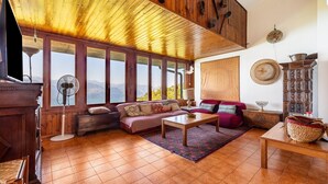 A cozy living room with a wooden ceiling, large windows, a sofa, a coffee table, and a traditional cabinet.