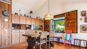 A wooden kitchen with a dining table and chairs, a hanging light fixture, and a window with a view of trees.