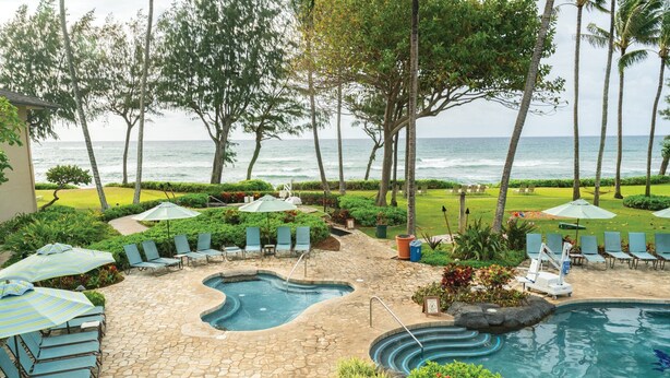 A pool area with lounge chairs and umbrellas, surrounded by greenery and palm trees, overlooking the ocean.