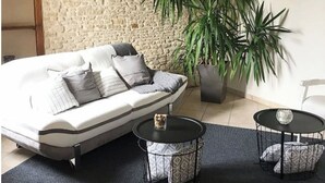Una habitación acogedora con pared de piedra, una planta grande en maceta, un sofá con cojines estampados y dos pequeñas mesas redondas con adornos.