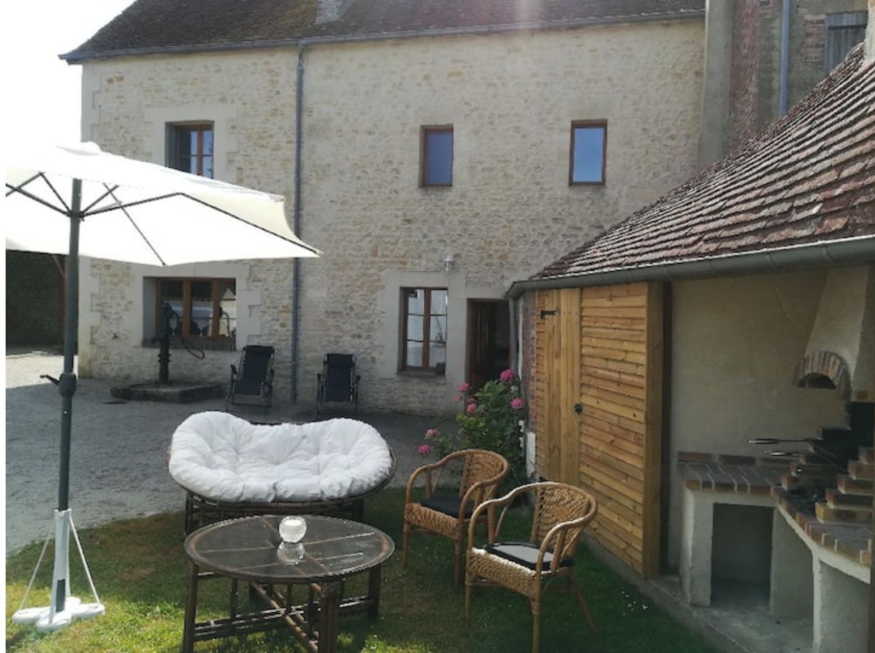 Un bâtiment en pierre avec un toit en tuiles, un espace extérieur abrité avec des chaises et une table, et une petite construction en bois dotée d’une cheminée.