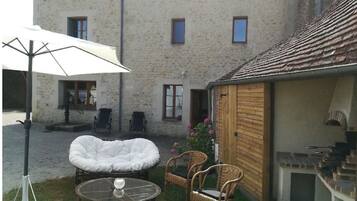 Un bâtiment en pierre avec un toit en tuiles, un espace extérieur abrité avec des chaises et une table, et une petite construction en bois dotée d’une cheminée.