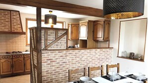 Cocina con armarios de madera, pared de ladrillo y zona de comedor preparada para cuatro personas, con manteles y platos blancos.