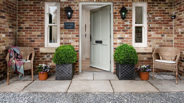 A brick building with a white door, two potted plants, and wicker chairs.