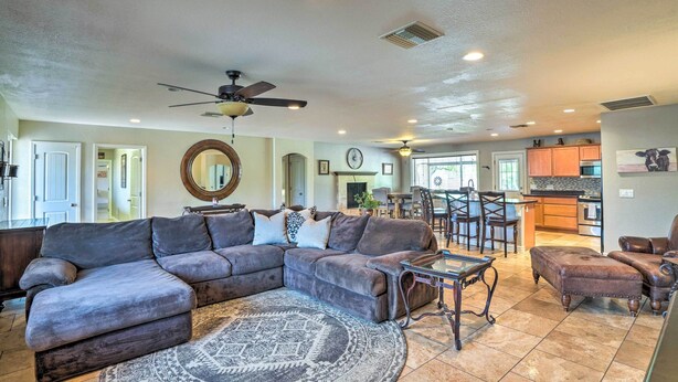 A spacious living room with a large sectional sofa, a round coffee table, and a patterned rug. There is a ceiling fan, a kitchen area in the background, and a dining area with bar stools.