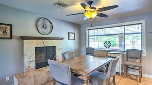 A dining room with a wooden table, six chairs, a fireplace, a ceiling fan, and a window with blinds.