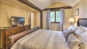 Une chambre avec une commode en bois, un téléviseur, un lit avec une literie à motifs, une fenêtre avec des rideaux et un ventilateur de plafond.
