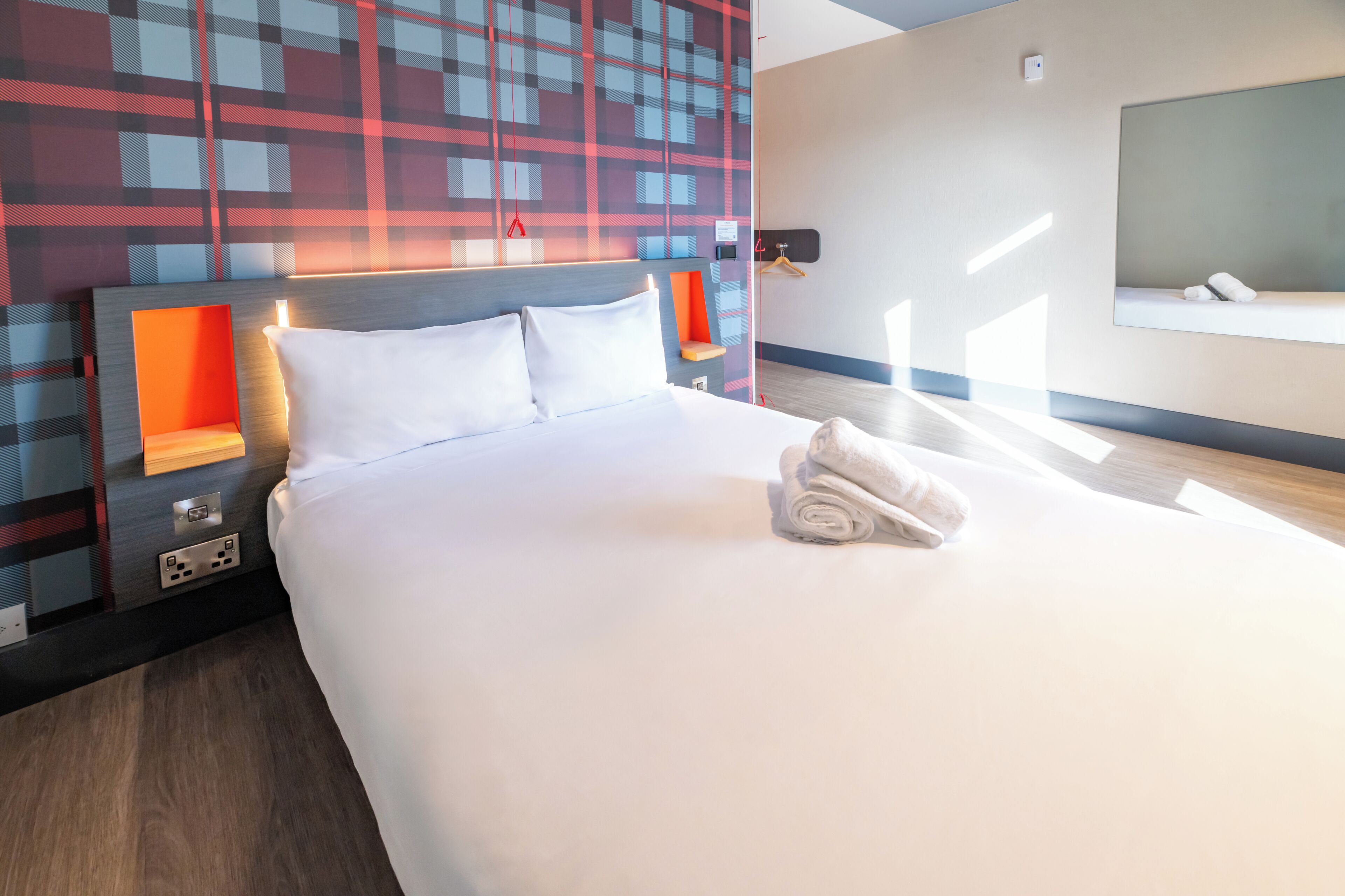 A hotel room with a bed, a headboard, a wall-mounted TV, and a window.