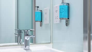 Un baño con un lavamanos blanco, grifo de cromo y espejo. Hay dos dispensadores en la pared, uno etiquetado 'SHAMERS ONLY' y otro 'SHAMERS ONLY' con etiqueta azul.