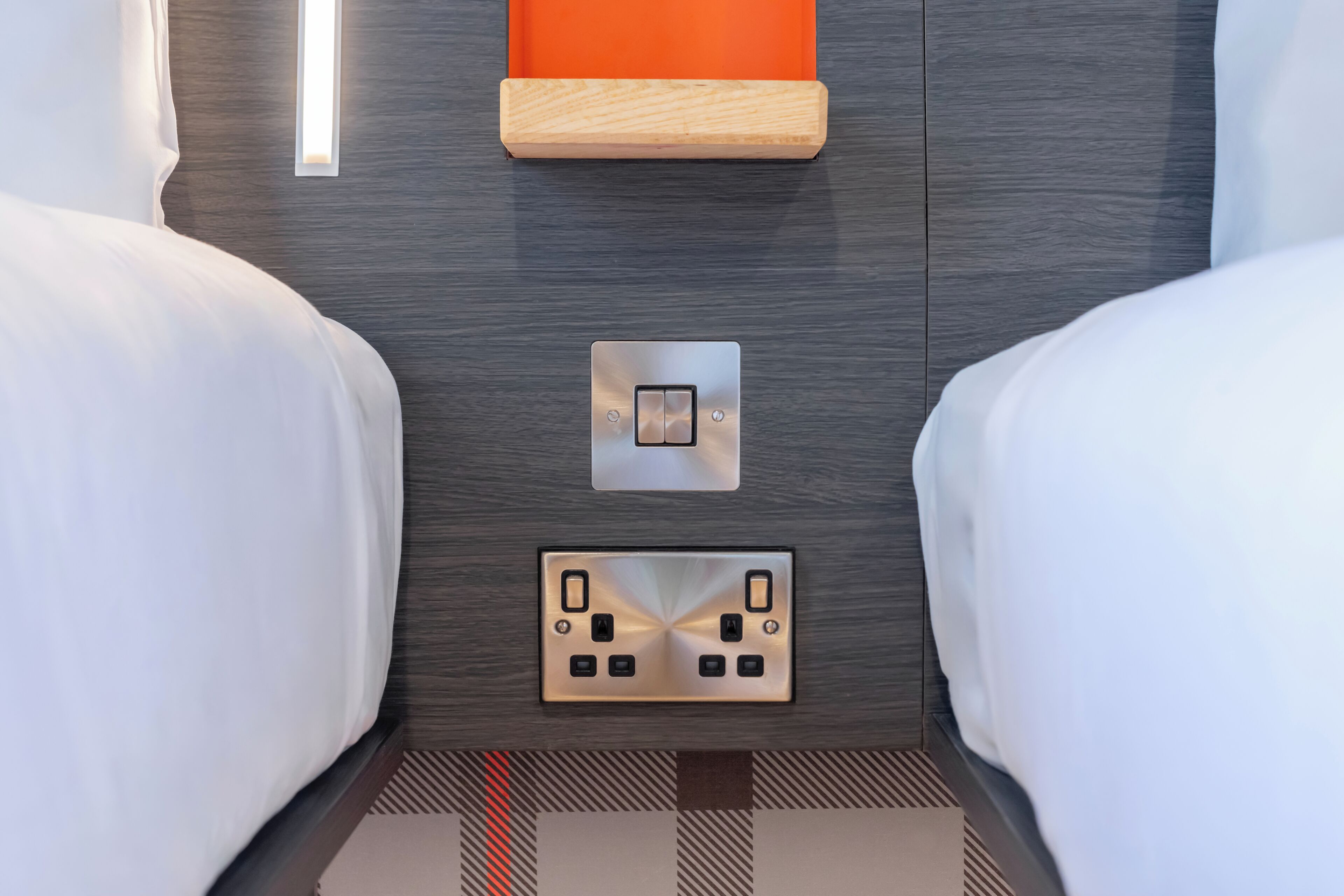A hotel room with two beds, a wall-mounted light, a power outlet, and a switch.