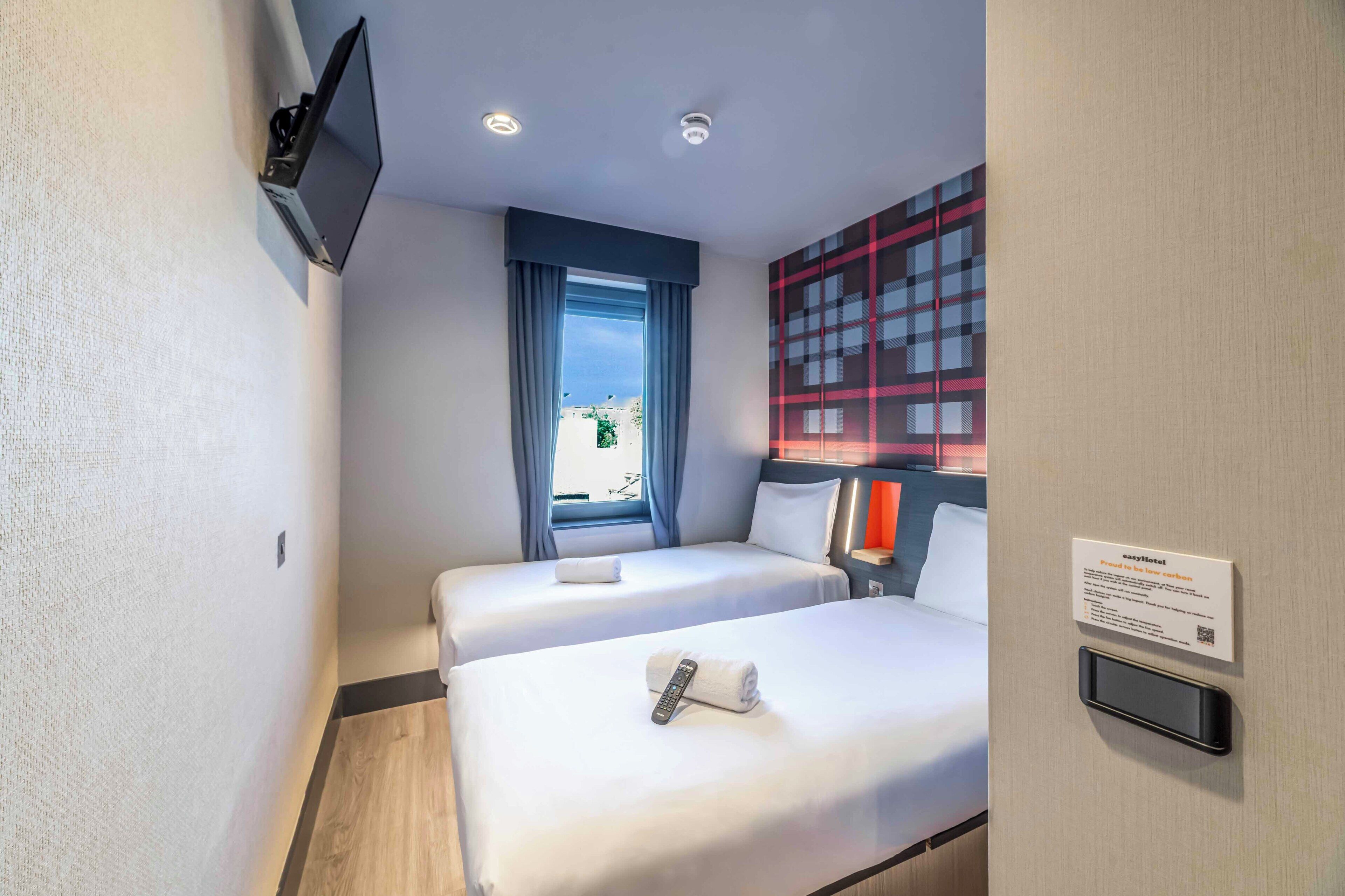 Habitación de hotel con dos camas, un televisor montado en la pared, una ventana con cortinas y un cartel con información.