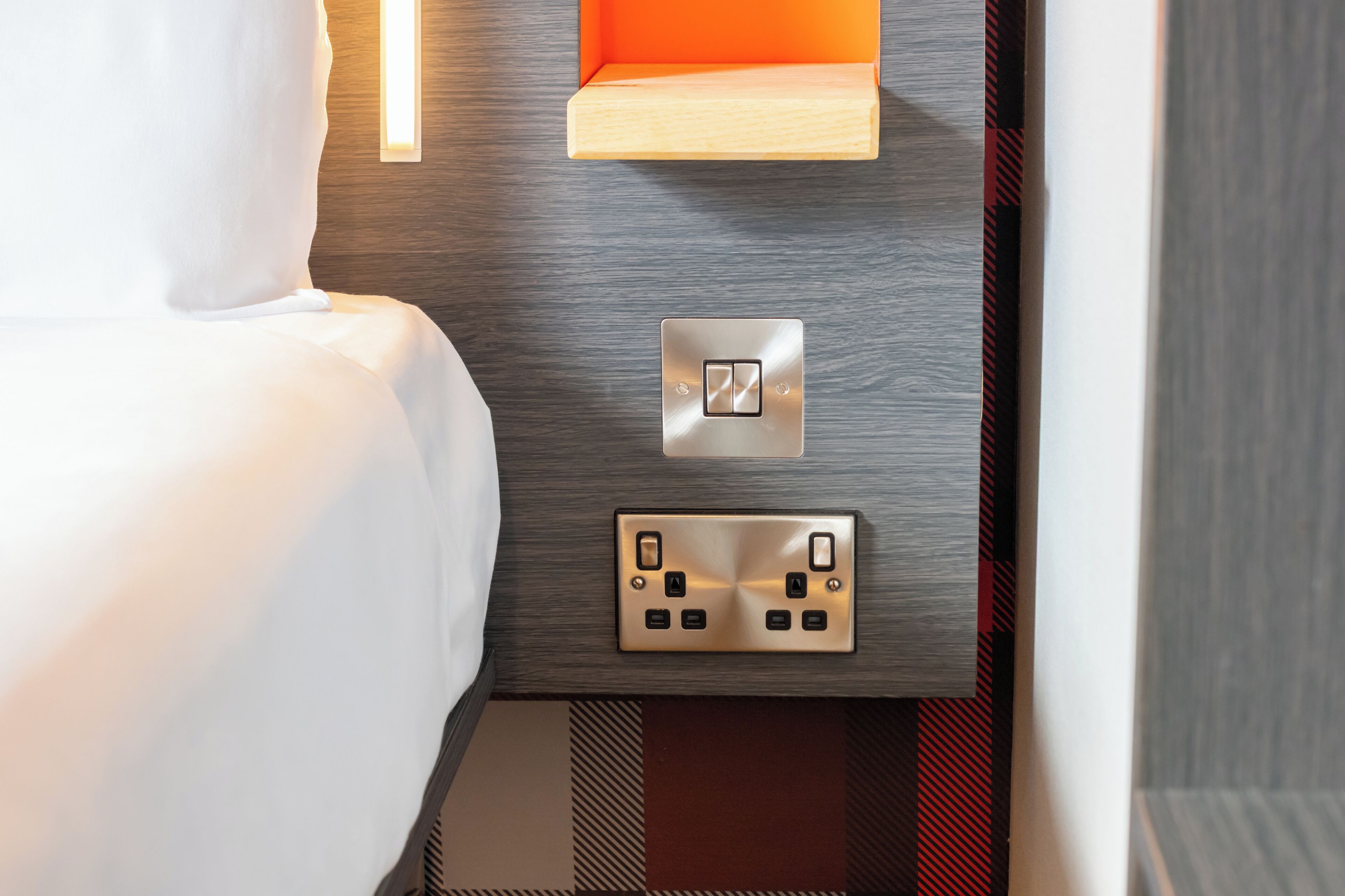 A hotel room with a bed, a wall-mounted light, a shelf, and a control panel.