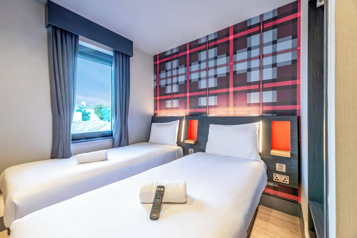 Ein Hotelzimmer mit zwei Betten, einem fensterbelaubten Fenster, einer Wand mit geometrischem Muster und einer Fernbedienung auf dem Bett.