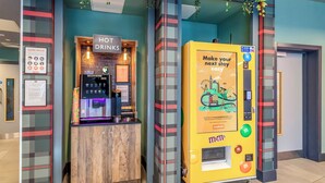 A hotel lobby with a 'HOT DRINKS' kiosk, a vending machine, and a 'HOTEL RULES' sign.