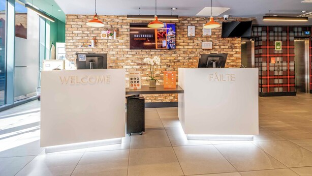 A reception area with a brick wall, a TV, and two counters labeled 'WELCOME' and 'FAILITE'.