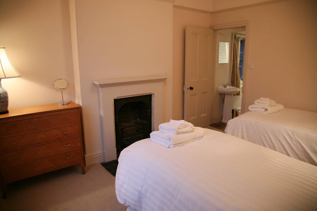 A hotel room with two beds, a wooden dresser, a mirror, a fireplace, and a bathroom with a sink and towel rack.
