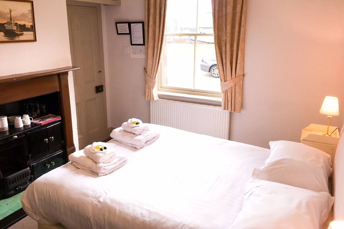 A hotel room with a bed, a nightstand with a lamp, a window with curtains, a radiator, a cabinet with towels, and a framed picture on the wall.