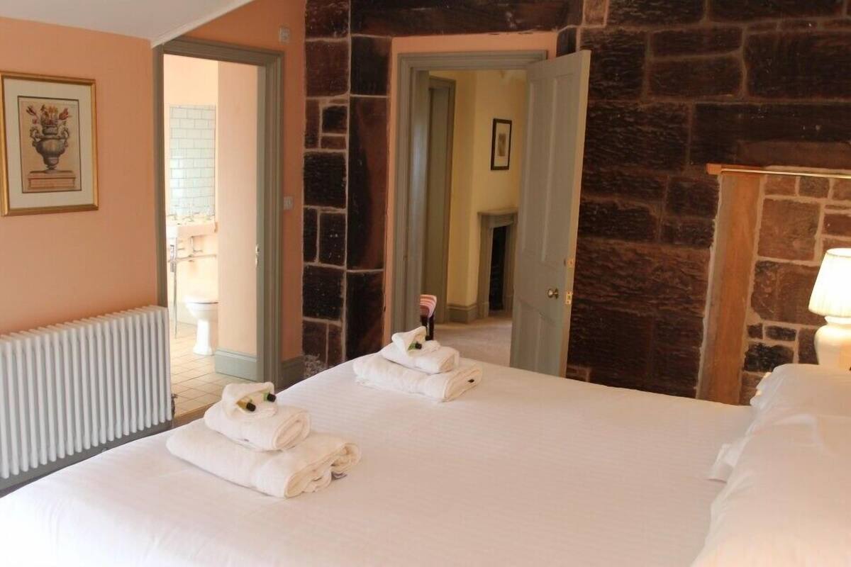 A hotel room with a large bed, two towels, a radiator, a stone wall, and a door leading to a bathroom.