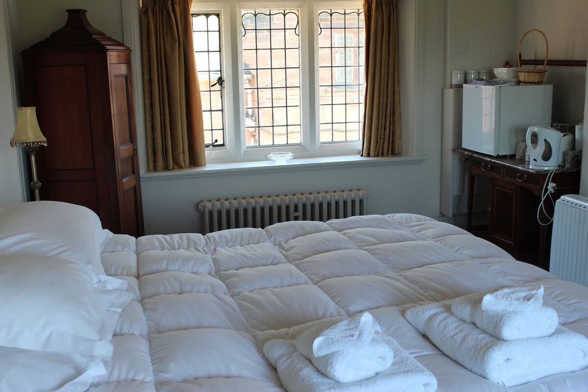 A hotel room with a large bed, a wooden wardrobe, a radiator, a window with curtains, and a kitchenette area.