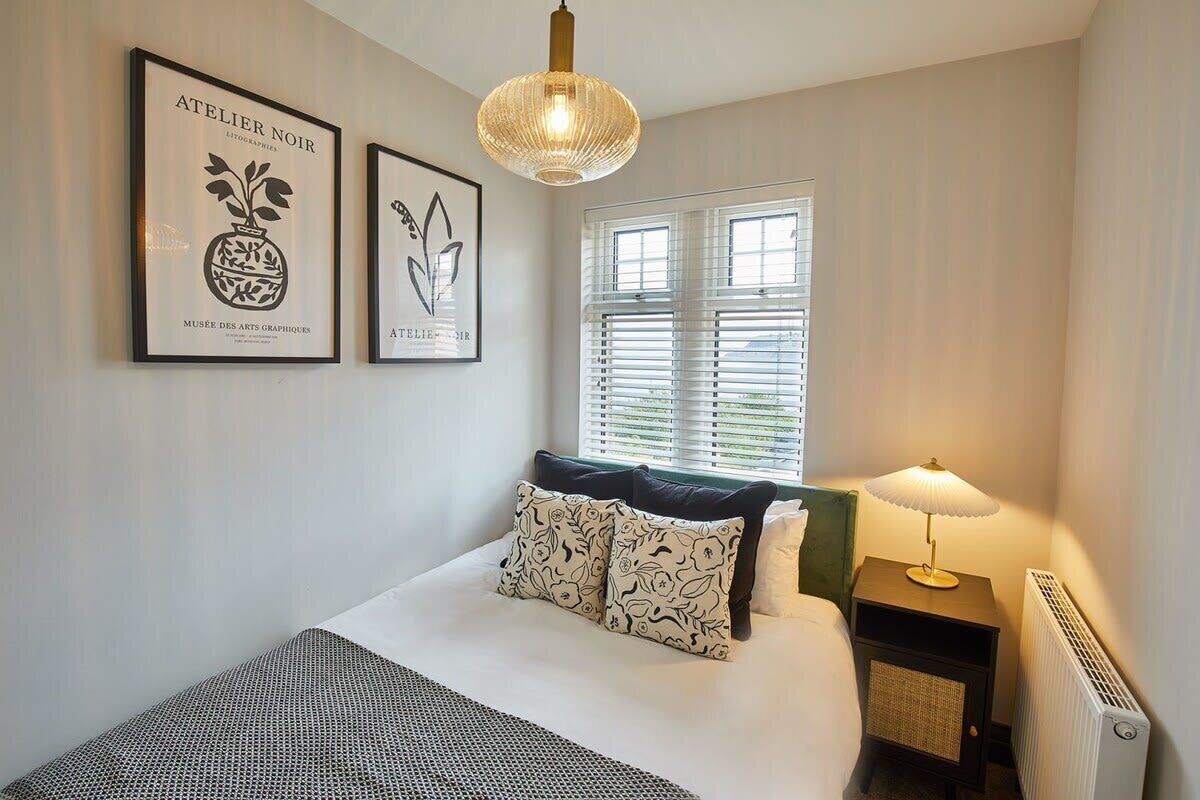 Une chambre à coucher avec un lit, deux œuvres d’art encadrées, une fenêtre avec des stores, une lampe de chevet et un radiateur.