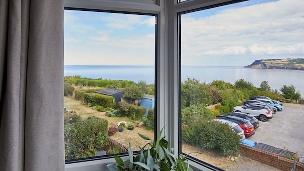 Une vue depuis une fenêtre donnant sur un parking où sont garées des voitures, ainsi que sur un paysage verdoyant avec une étendue d’eau et des collines à l’horizon.