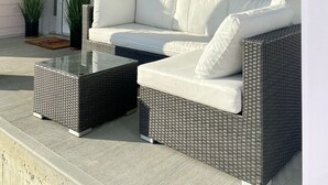 A covered porch with a white wicker sofa and two matching ottomans, a glass coffee table, and potted plants. - 2026-27 October to May deep discount special pricing! Please contact for rates! (Port Sranley)