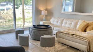 A modern living room with a white leather sofa, grey ottomans, a round coffee table, and a large window offering a view of a construction site. - 2026-27 October to May deep discount special pricing! Please contact for rates! (Port Sranley)