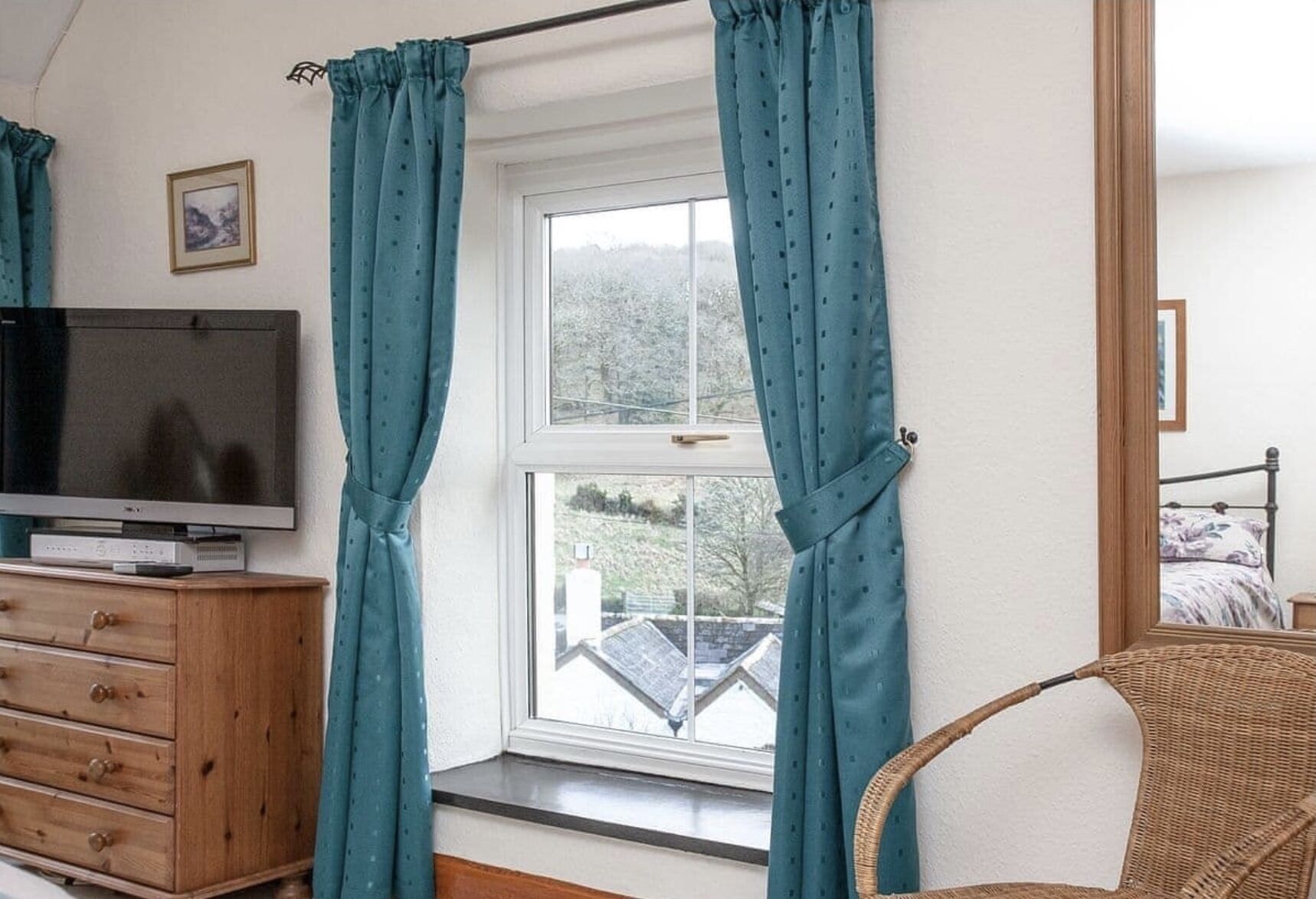 Ein Zimmer mit Fernseher auf einer Holzkommode, einem Fenster mit türkisfarbenen Vorhängen, einem Korbstuhl und einem Bett mit floralem Überwurf.