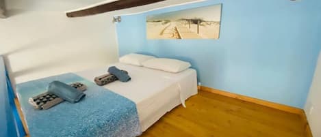 A small, single-bed room with a wooden floor, a blue wall, and a beach-themed picture.